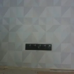 Ściana pokryta tapetą w geometryczne, trójwymiarowe wzory w odcieniach szarości, z zamontowanym czarnym panelem gniazdek i włączników.