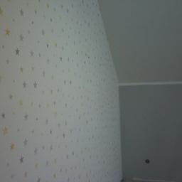 SW Projekt - Ściana w pokoju na poddaszu pokryta tapetą w białe tło z szarymi i żółtymi gwiazdkami, widoczny fragment drugiej ściany pomalowanej na szaro oraz białe wiadro z farbą.