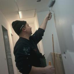 SW Projekt - Mężczyzna w czapce maluje ścianę wałkiem w trakcie remontu korytarza, widoczne niedomalowane fragmenty i przygotowane narzędzia.