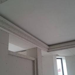 SW Projekt - Wnętrze w trakcie remontu z widoczną konstrukcją sufitu podwieszanego z płyt gipsowo-kartonowych, obejmującą także fragment ściany, z widocznymi otworami na śruby, oraz okna w tle.