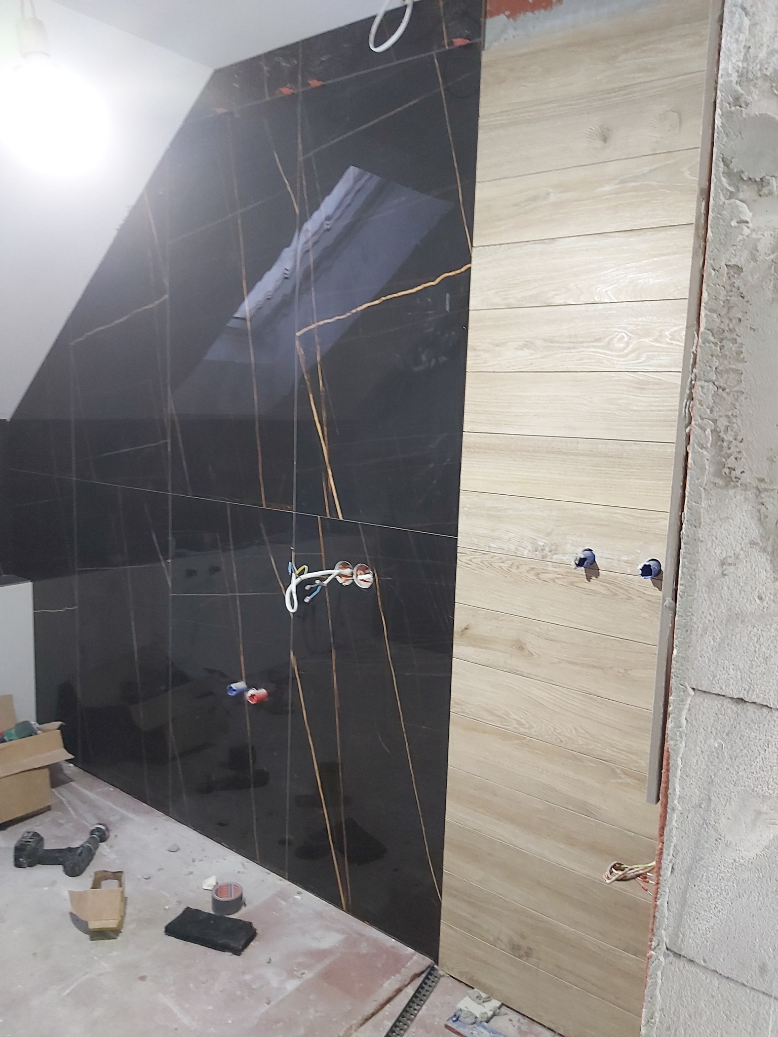 Częściowo wykończona ściana łazienki z czarnymi płytkami z złotymi żyłami i drewnopodobnymi panelami; widoczne wystające kable i narzędzia na podłodze w trakcie prac.