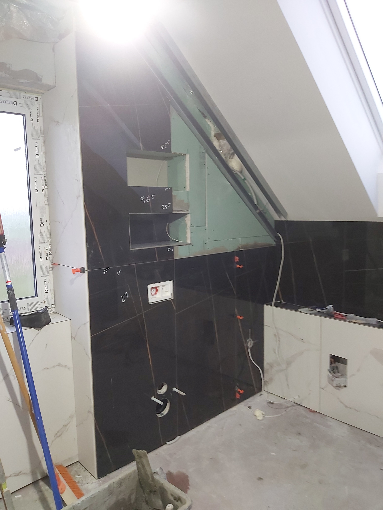 Montaż czarnych płytek w łazience na poddaszu, widoczne przycięte płytki, poziomica, elementy instalacji i przymocowane krzyżyki dystansowe.