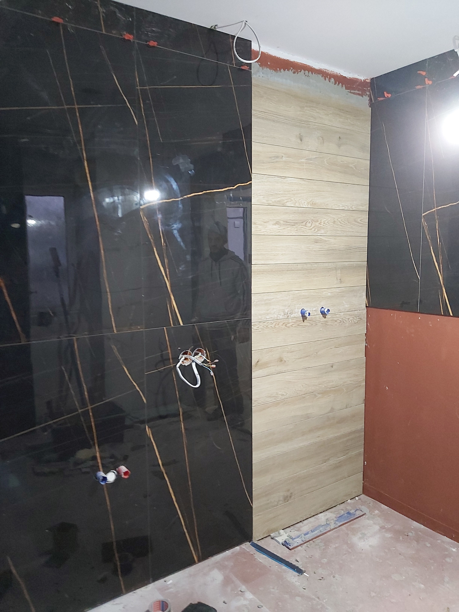 Częściowo wykończona łazienka z czarnymi płytkami imitującymi marmur z złotymi żyłami na ścianie i podłodze, sąsiadującymi z jasnymi płytkami imitującymi drewno na ścianie, widoczne wystające kable...