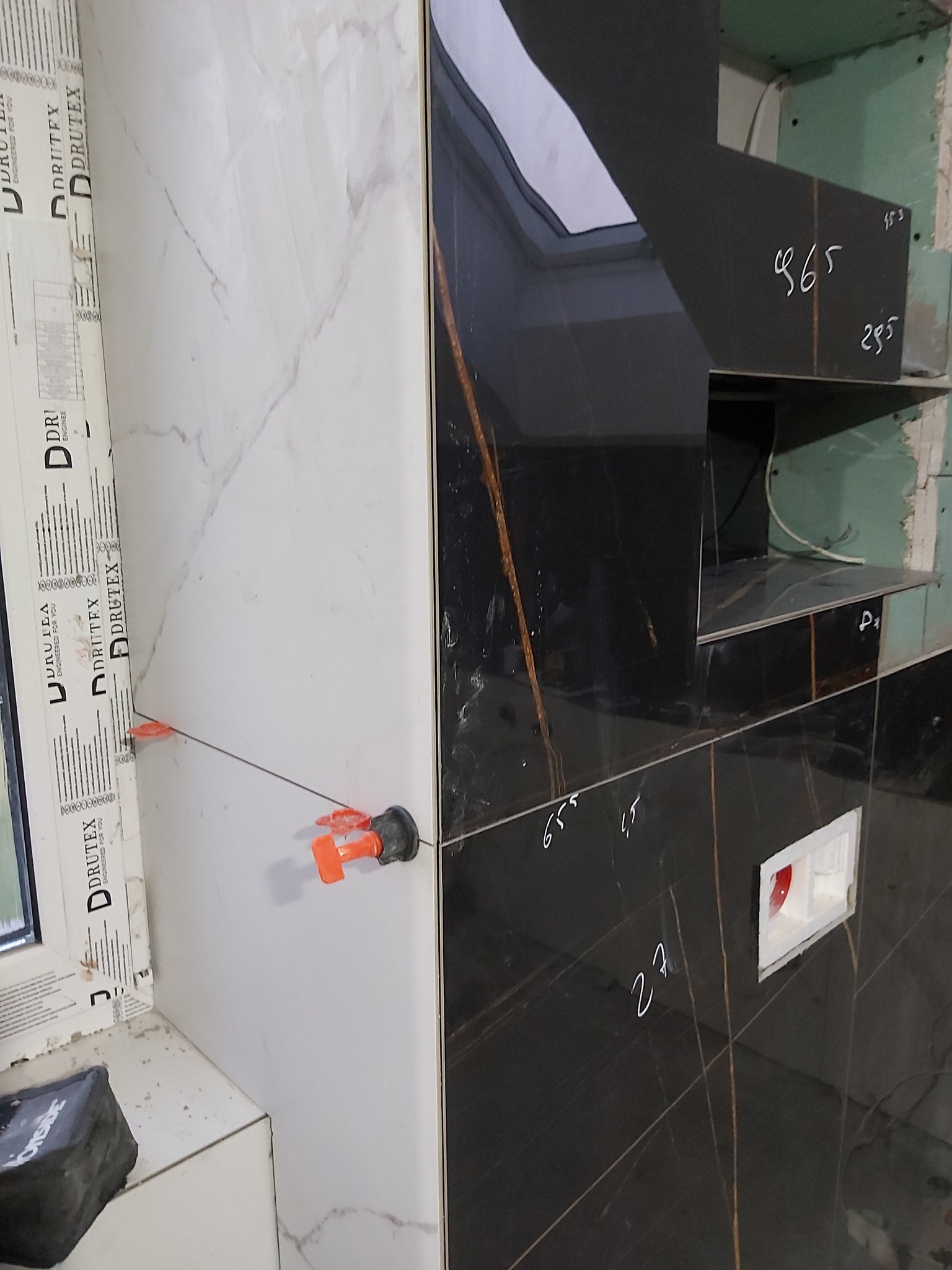 Białe i czarne płytki imitujące marmur podczas układania w łazience, z użyciem systemu poziomowania płytek i zapiskami ołówkiem na czarnych płytkach.