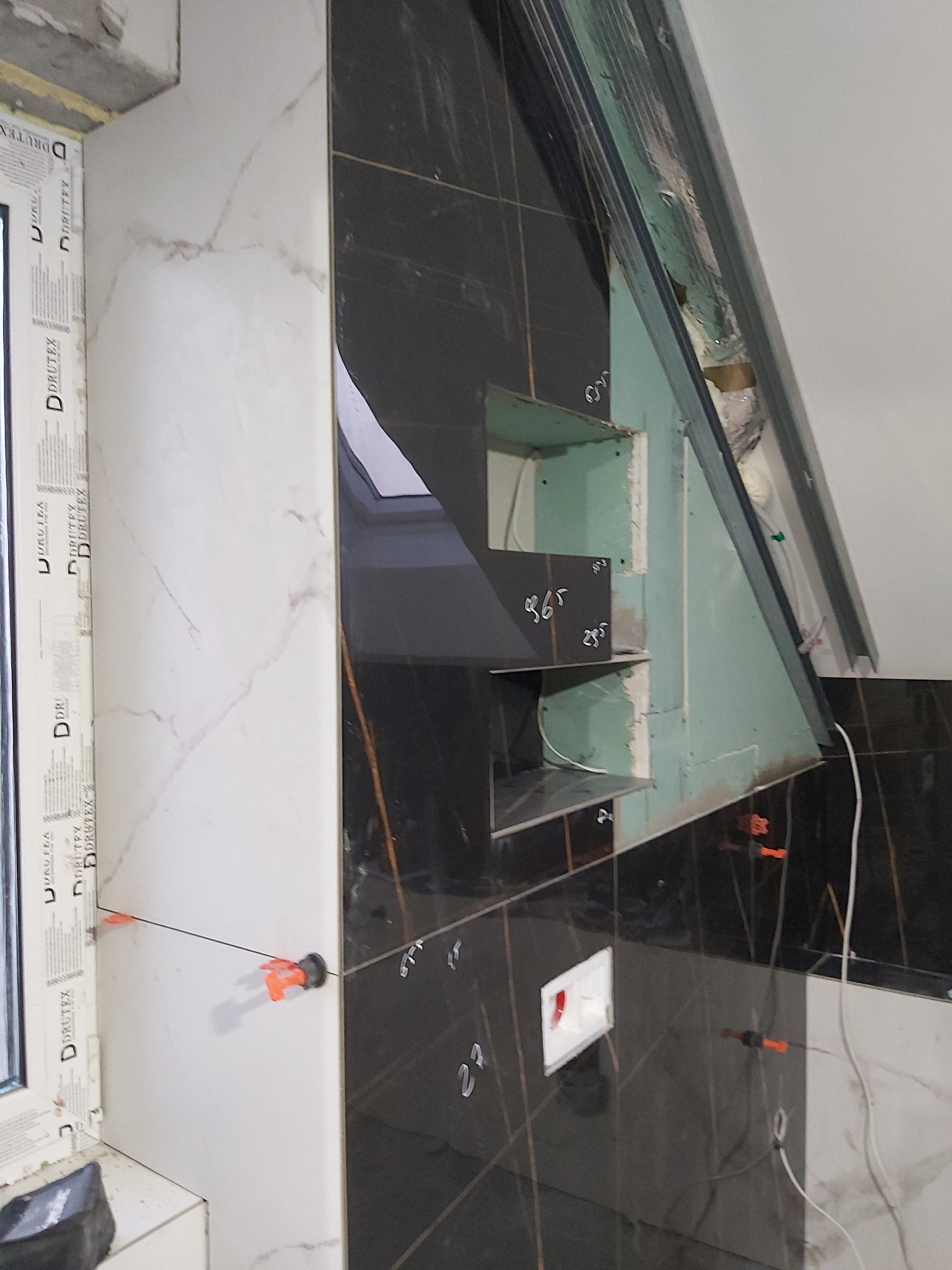 Układanie czarnych płytek w łazience, widoczne poziomice i krzyżyki dystansowe, półki z płyty gipsowej w trakcie wykańczania, okno z widocznym napisem 'DRUTEX'.
