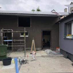 Grzeskowiak - Elewacja budynku po nałożeniu tynku maszynowego, widoczne rusztowanie, narzędzia i worki z zaprawą, przygotowane do dalszych prac wykończeniowych.