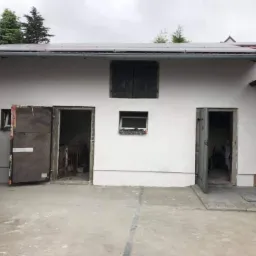 Biała fasada budynku gospodarczego z otwartymi drzwiami i oknem z okiennicami, widoczne panele słoneczne na dachu.