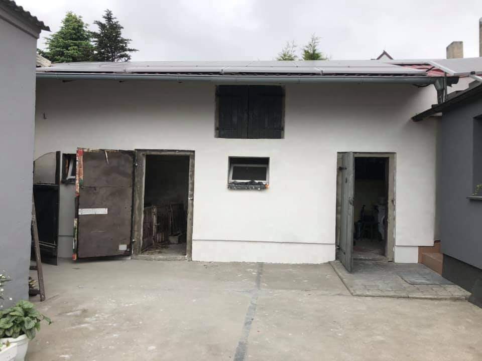 Biała fasada budynku gospodarczego z otwartymi drzwiami i oknem z okiennicami, widoczne panele słoneczne na dachu.