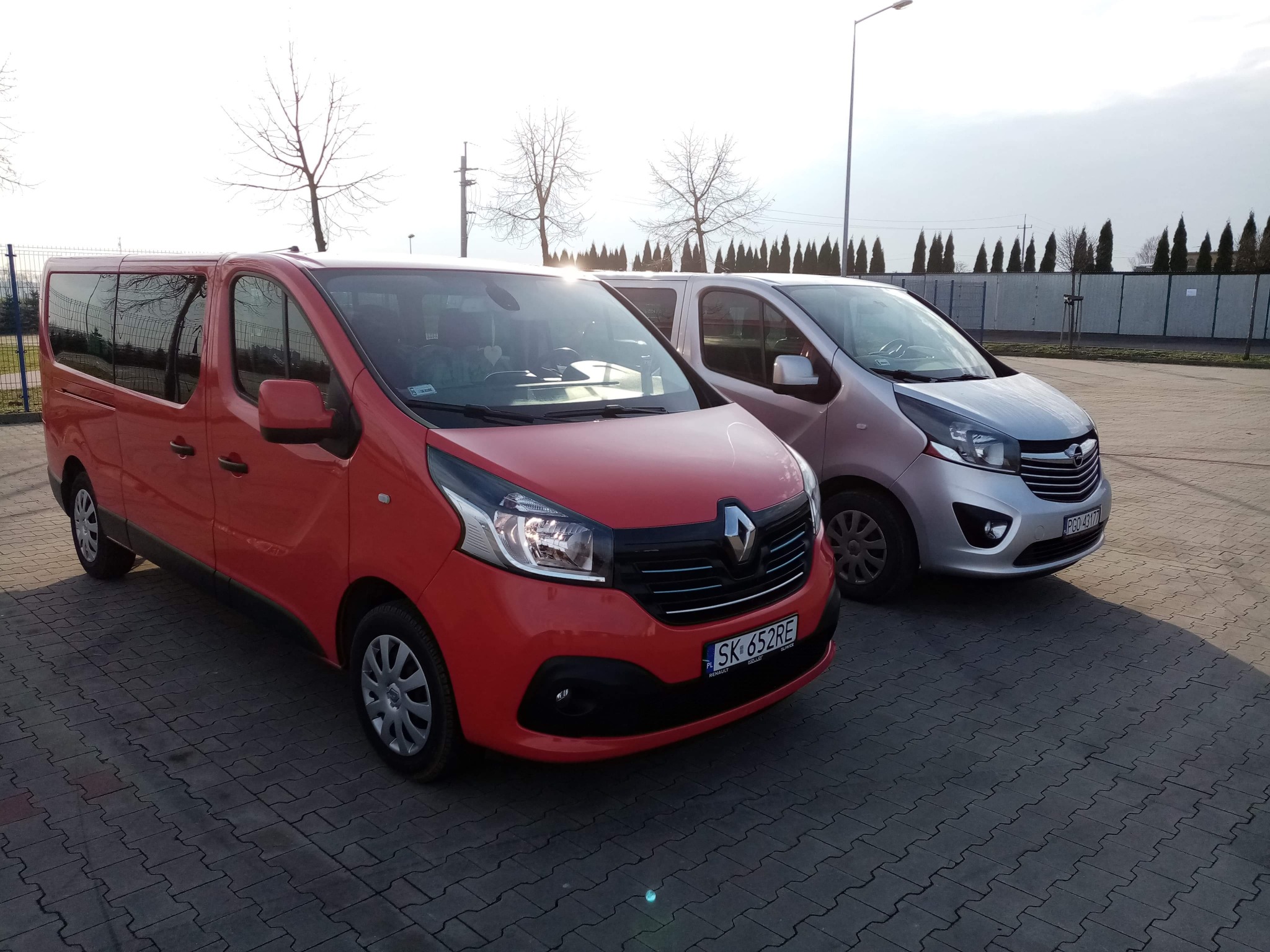 Dwa busy: czerwony Renault Trafic i srebrny Opel Vivaro zaparkowane na brukowanym placu w słoneczny dzień, z widocznym ogrodzeniem i drzewami w tle.
