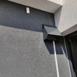 Szary anemostat zewnętrzny z kratką i białą rurą wentylacyjną na elewacji budynku o szarej, chropowatej strukturze, obok fragmentu czarnej ramy okiennej.