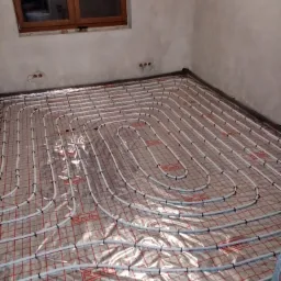 Białe rury systemu grzewczego ułożone spiralnie na czerwonej siatce pod folią w surowym pomieszczeniu z widocznym oknem i wystającymi kablami elektrycznymi.