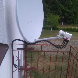 Biała antena satelitarna zamontowana na metalowej balustradzie balkonu, z widocznymi kablami i złączami, na tle ściany budynku i drzew.