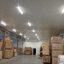 Wnętrze hali magazynowej z oświetleniem LED, instalacją elektryczną w korytkach i nagrzewnicą powietrza, palety z drewnem składowane wzdłuż ścian.