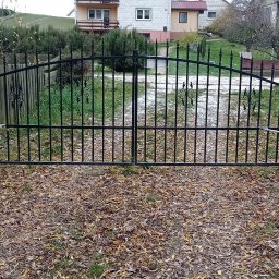 MS-INSTAL Marcin Sudoł - Czarna, metalowa brama dwuskrzydłowa z automatyką zamontowaną na betonowych słupkach, otoczona opadłymi liśćmi.