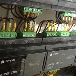 Szereg modułów przekaźnikowych z zielonymi złączkami, oznaczonymi przewodami i żółtymi etykietami, zamontowanych w szafie sterowniczej z widocznymi przewodami.