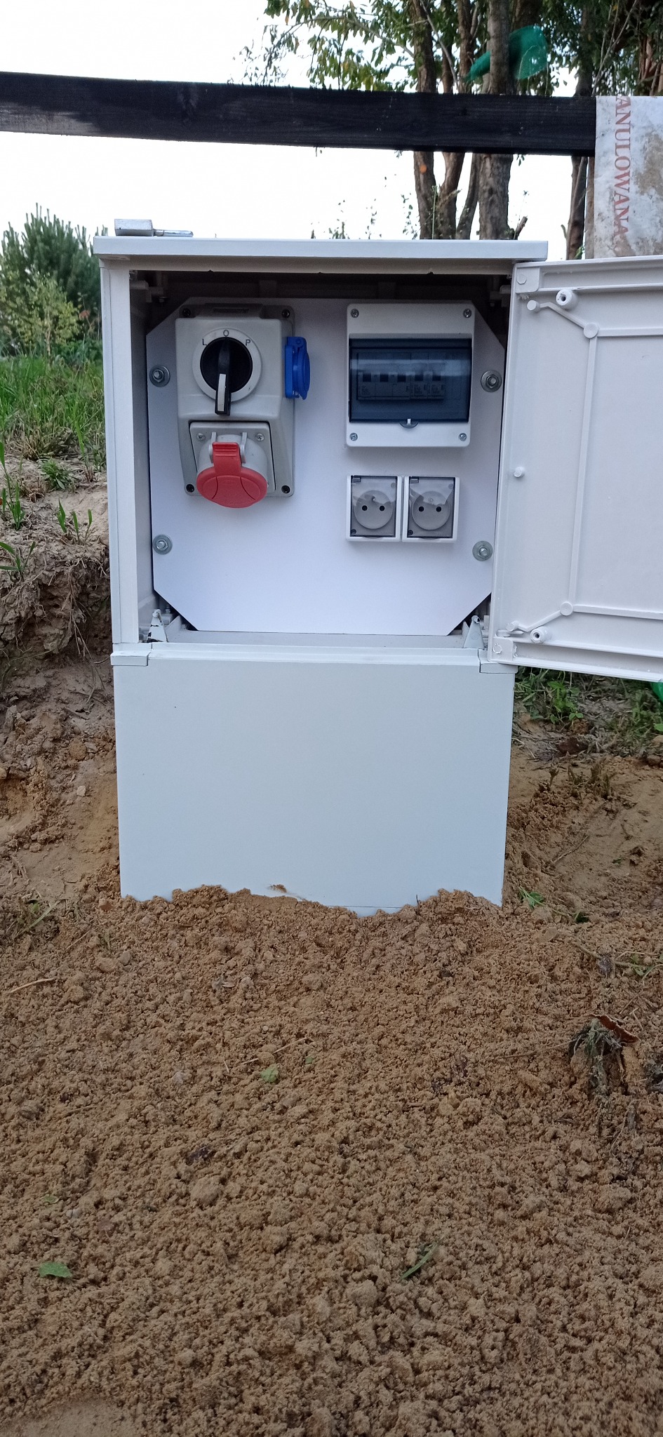 Biała skrzynka elektryczna z otwartymi drzwiczkami, zawierająca gniazda, wyłącznik i zabezpieczenia, zamontowana w ziemi na zewnątrz.