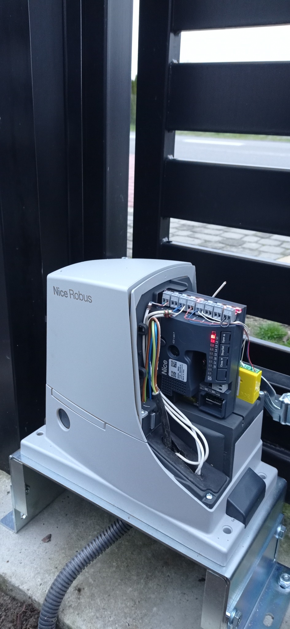 Szary automat do bramy Nice Robus z widocznym panelem sterowania, okablowaniem i diodami LED, zamontowany przy czarnej bramie.