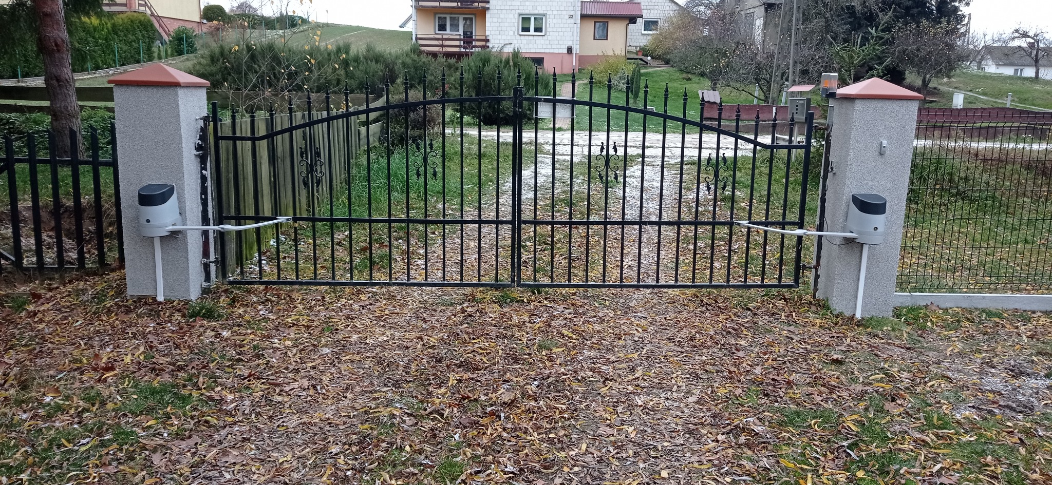 Czarna, metalowa brama dwuskrzydłowa z automatyką zamontowaną na betonowych słupkach, otoczona opadłymi liśćmi.