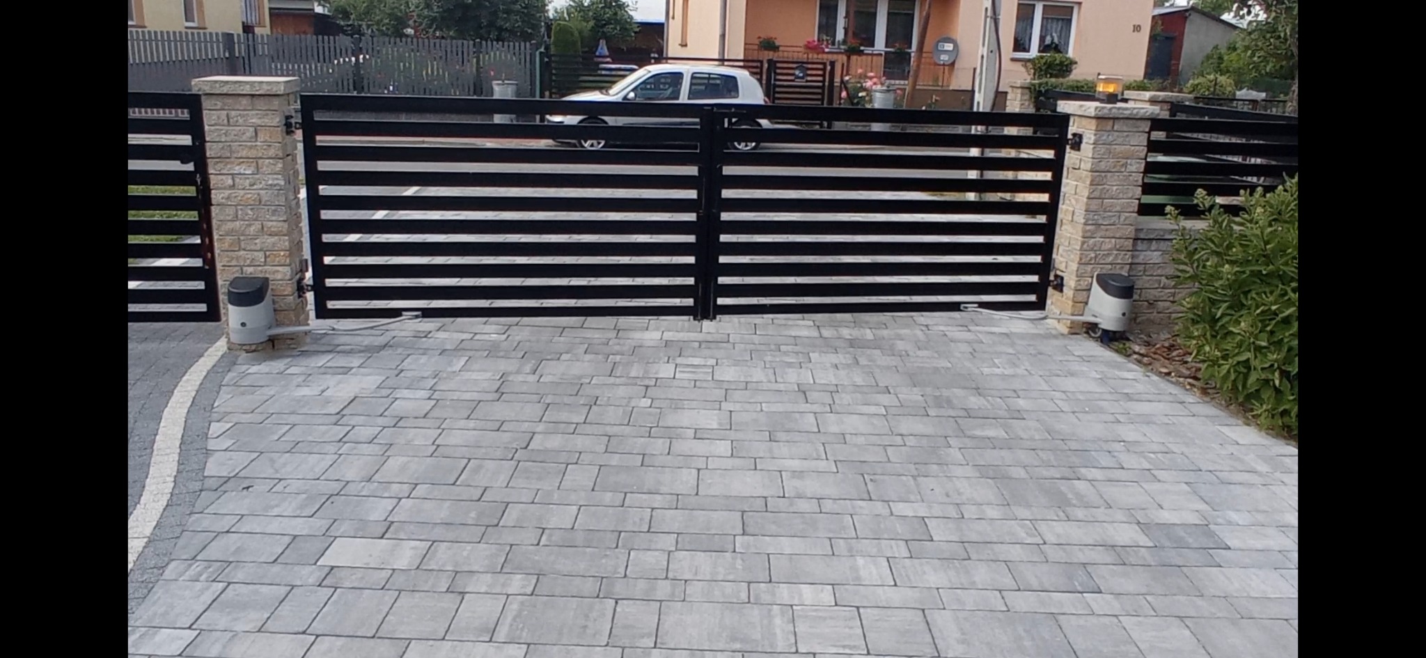 Nowoczesna brama wjazdowa z automatyką, czarne poziome panele, kamienne słupki, szara kostka brukowa na podjeździe, widoczny napęd bramy.