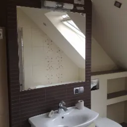 Ukończona łazienka na poddaszu z oknem dachowym, beżowe i brązowe płytki, umywalka z baterią, lustro z oświetleniem, toaleta, półki w ścianie, na podłodze stoi karton z płytkami Cersanit.