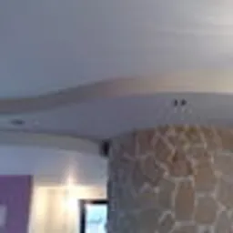 Częściowe ujęcie wnętrza z kolumną obłożoną kamieniem dekoracyjnym, widoczny fragment sufitu z ozdobnym wykończeniem i oświetleniem punktowym oraz ściana pomalowana na fioletowo.