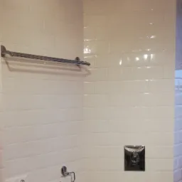 Ściana łazienki wyłożona białymi, błyszczącymi płytkami typu metro, z chromowanym wieszakiem na ręczniki, uchwytem na papier toaletowy i przyciskiem spłuczki w kwadratowej obudowie.