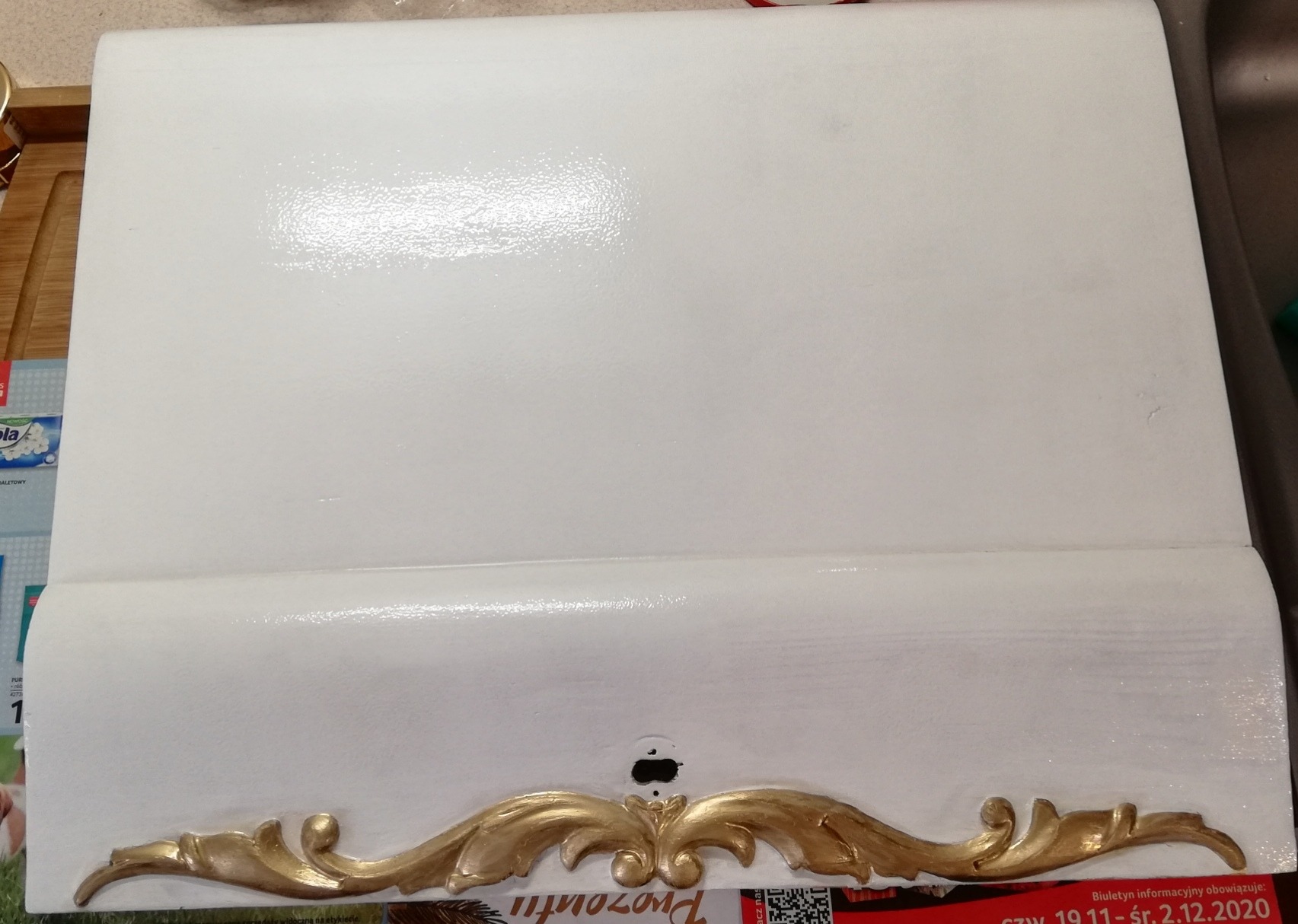 Świeżo pomalowana na biało szafka z dekoracyjnym, złotym ornamentem u dołu, widoczne ślady pędzla i połysk farby.