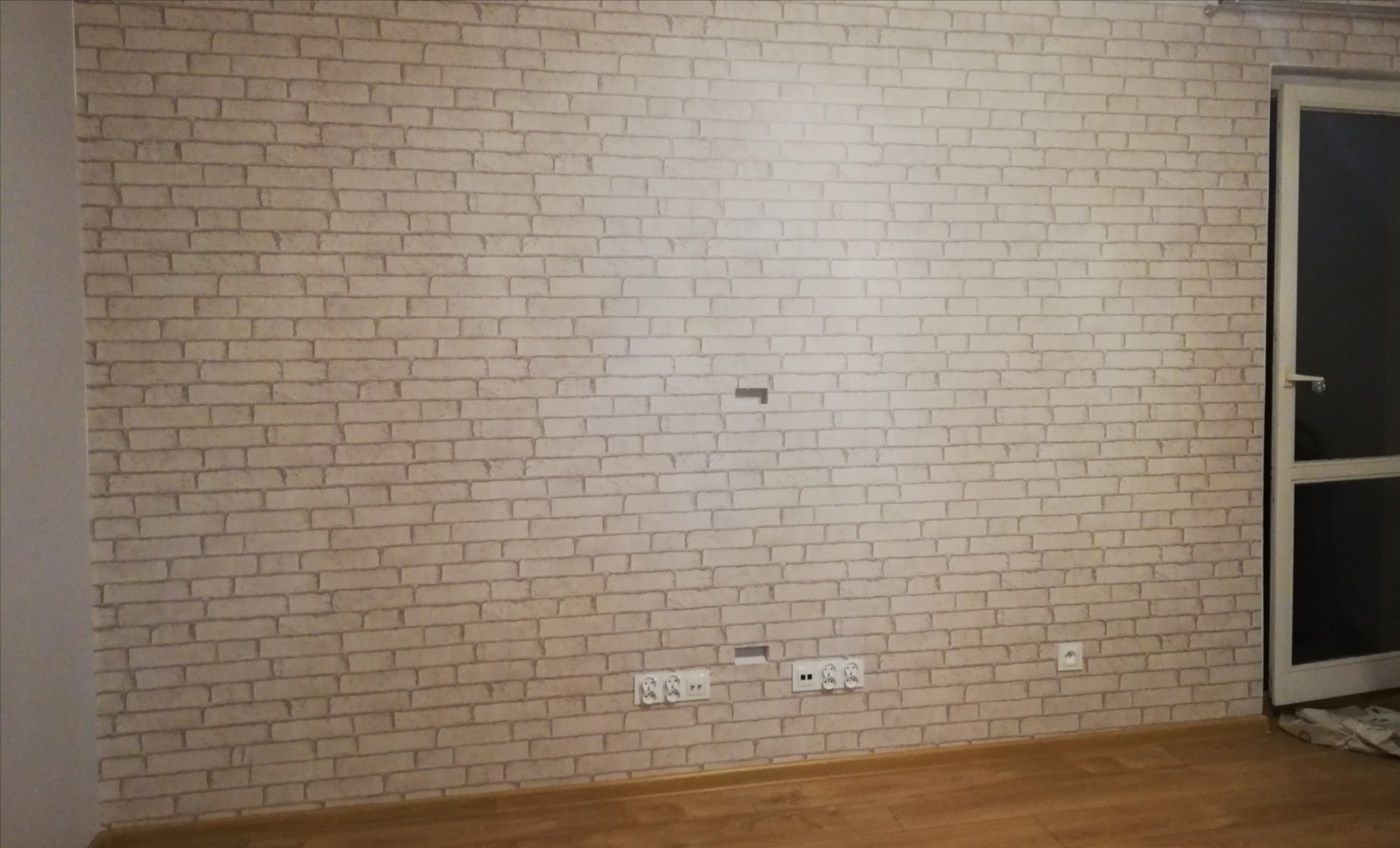 Tapeta imitująca białą cegłę na ścianie z widocznymi gniazdkami elektrycznymi i otworem na przewody, obok drzwi balkonowe.
