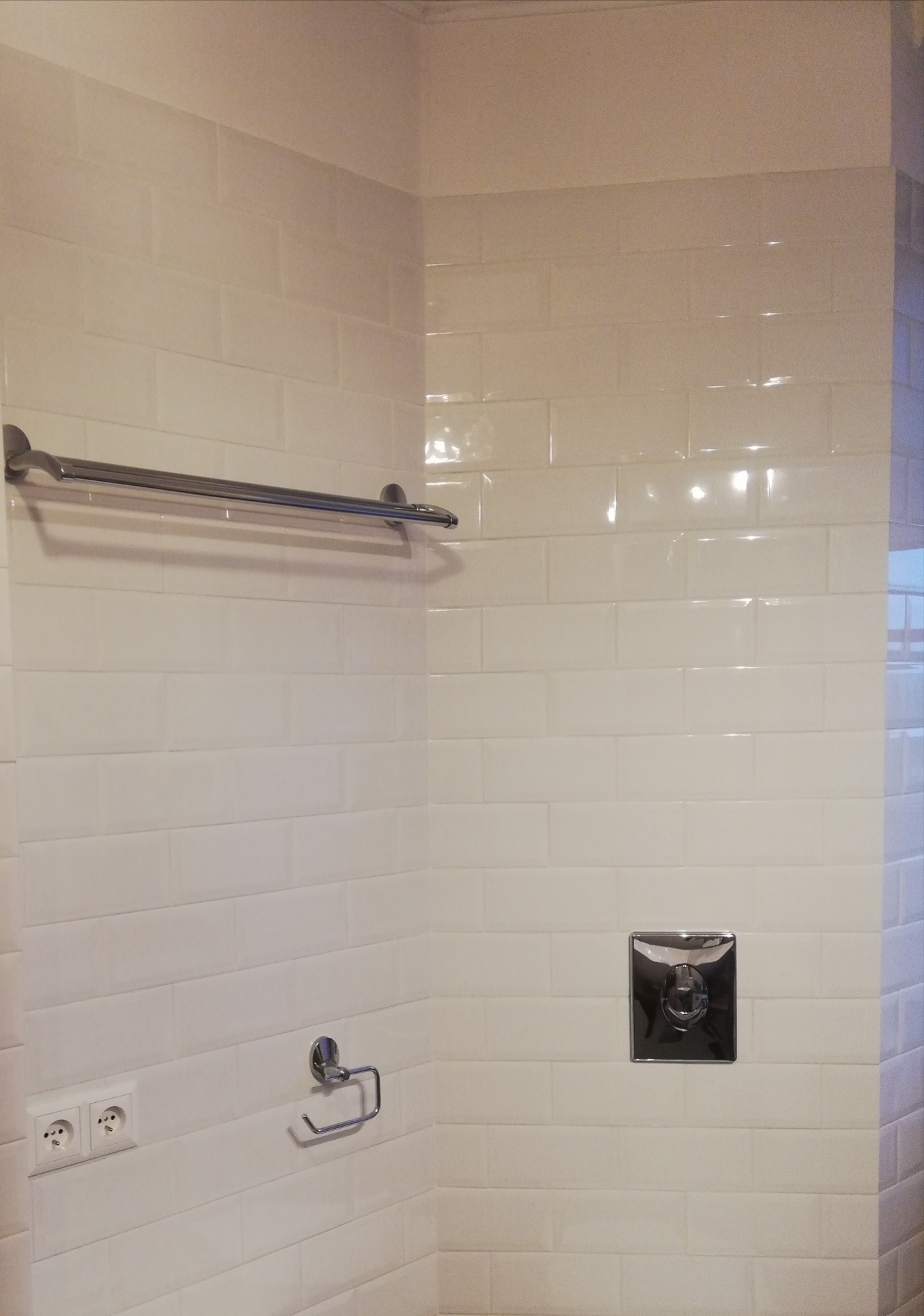 Ściana łazienki wyłożona białymi, błyszczącymi płytkami typu metro, z chromowanym wieszakiem na ręczniki, uchwytem na papier toaletowy i przyciskiem spłuczki w kwadratowej obudowie.