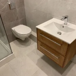 Nowoczesna łazienka z kabiną prysznicową, podwieszaną toaletą i drewnianą szafką z umywalką, wykończona płytkami imitującymi marmur.