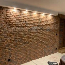 Ściana wykończona czerwoną cegłą klinkierową z widocznymi fugami, oświetlona punktowymi lampami sufitowymi, z drewnianymi drzwiami w tle.