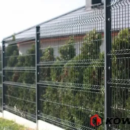 Ciemnoszare, nowoczesne ogrodzenie z paneli zgrzewanych na tle zielonych krzewów i domu jednorodzinnego.