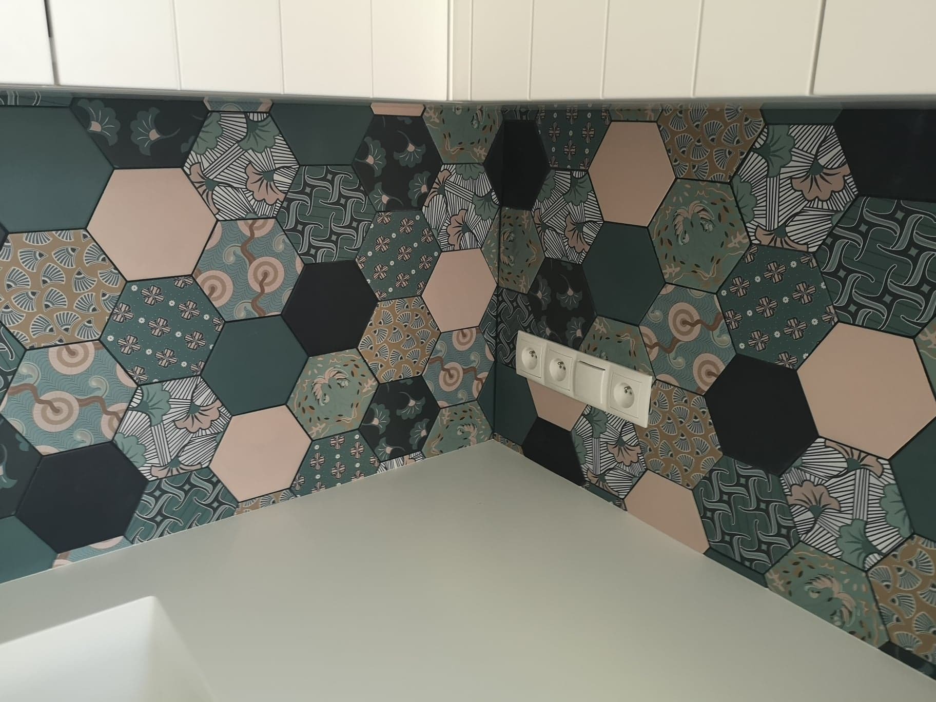 Ściana w kuchni wyłożona heksagonalnymi płytkami ceramicznymi w odcieniach zieleni, beżu i czerni z różnorodnymi wzorami, z widocznymi gniazdkami elektrycznymi i fragmentem białego blatu.