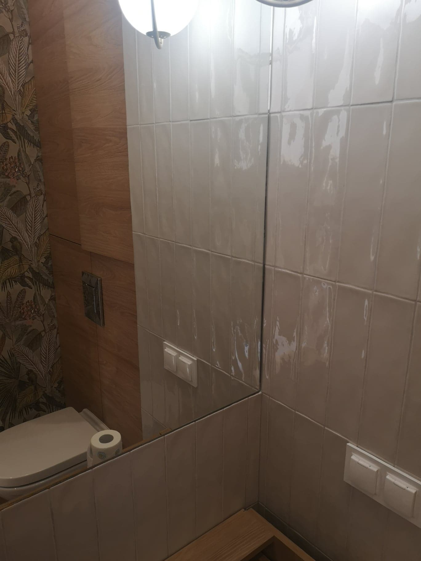 Ukończona łazienka z beżowymi, błyszczącymi płytkami ułożonymi pionowo, drewnianą zabudową i toaletą z rolką papieru toaletowego, widoczna w lustrze.