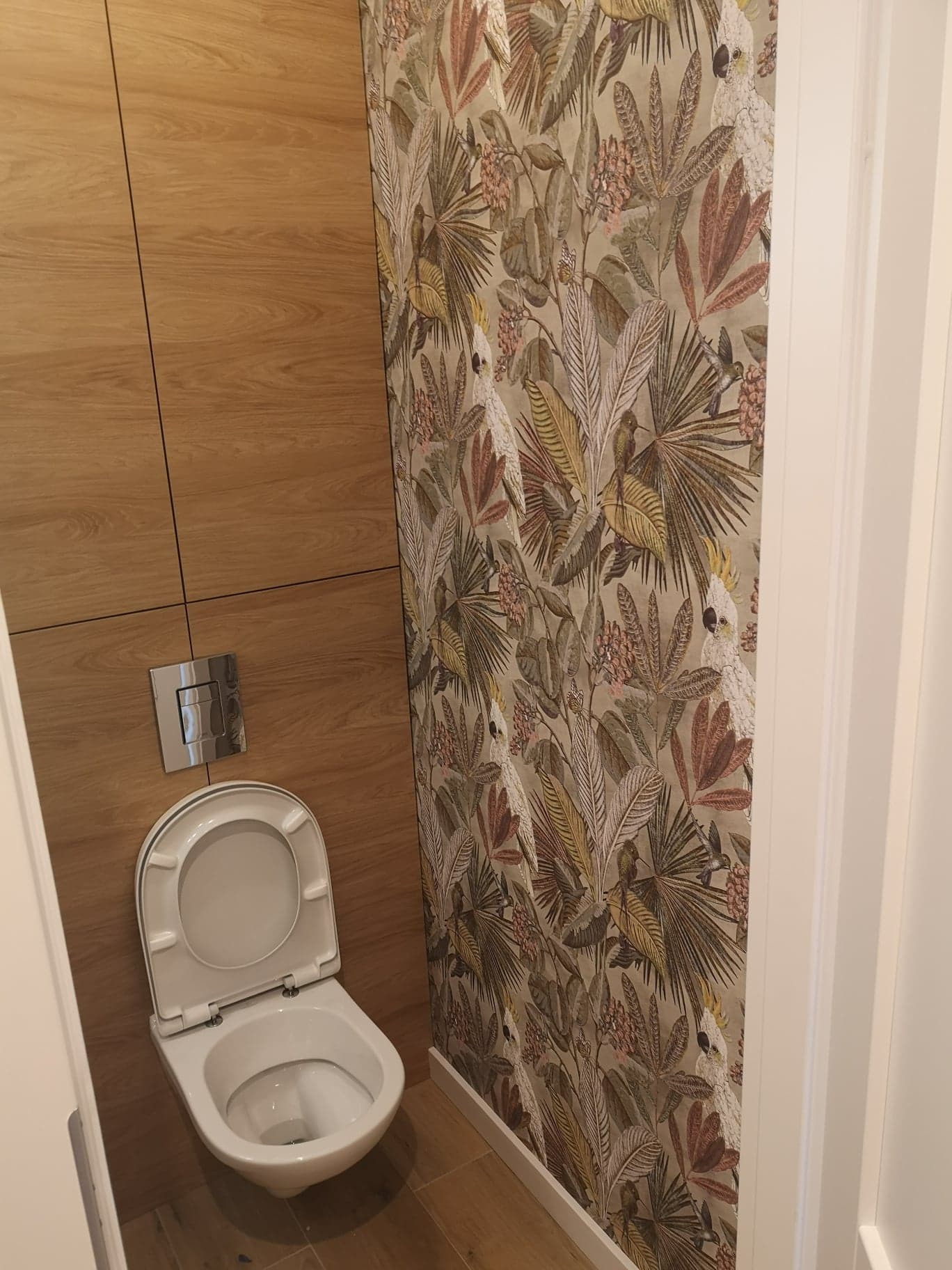 Toaleta w łazience z drewnopodobnymi płytkami i tapetą w tropikalne liście i papugi.