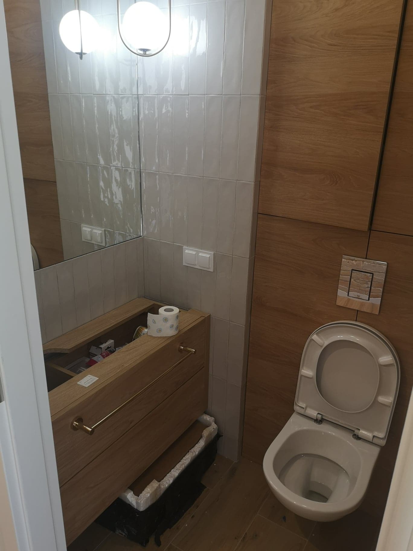 Nowoczesna łazienka w trakcie aranżacji: drewniane panele ścienne, białe płytki typu cegiełka, wisząca szafka z otwartymi drzwiczkami i złotymi uchwytami, toaleta z otwartą klapą, lustro...