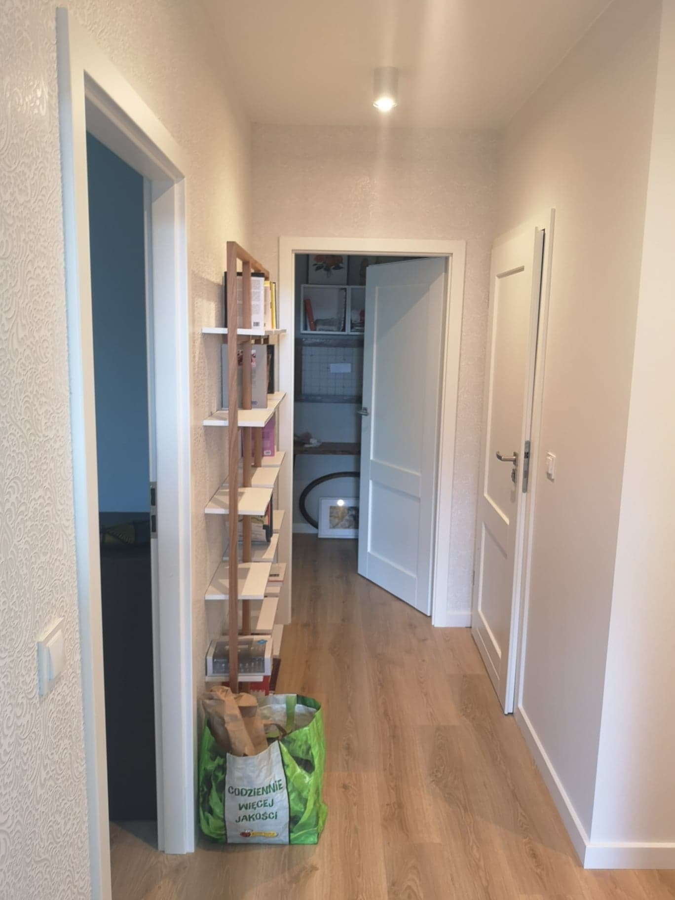 Wnętrze korytarza z drewnianą podłogą, białymi ścianami z delikatnym wzorem, regałem na książki i otwartymi drzwiami do pomieszczenia gospodarczego.
