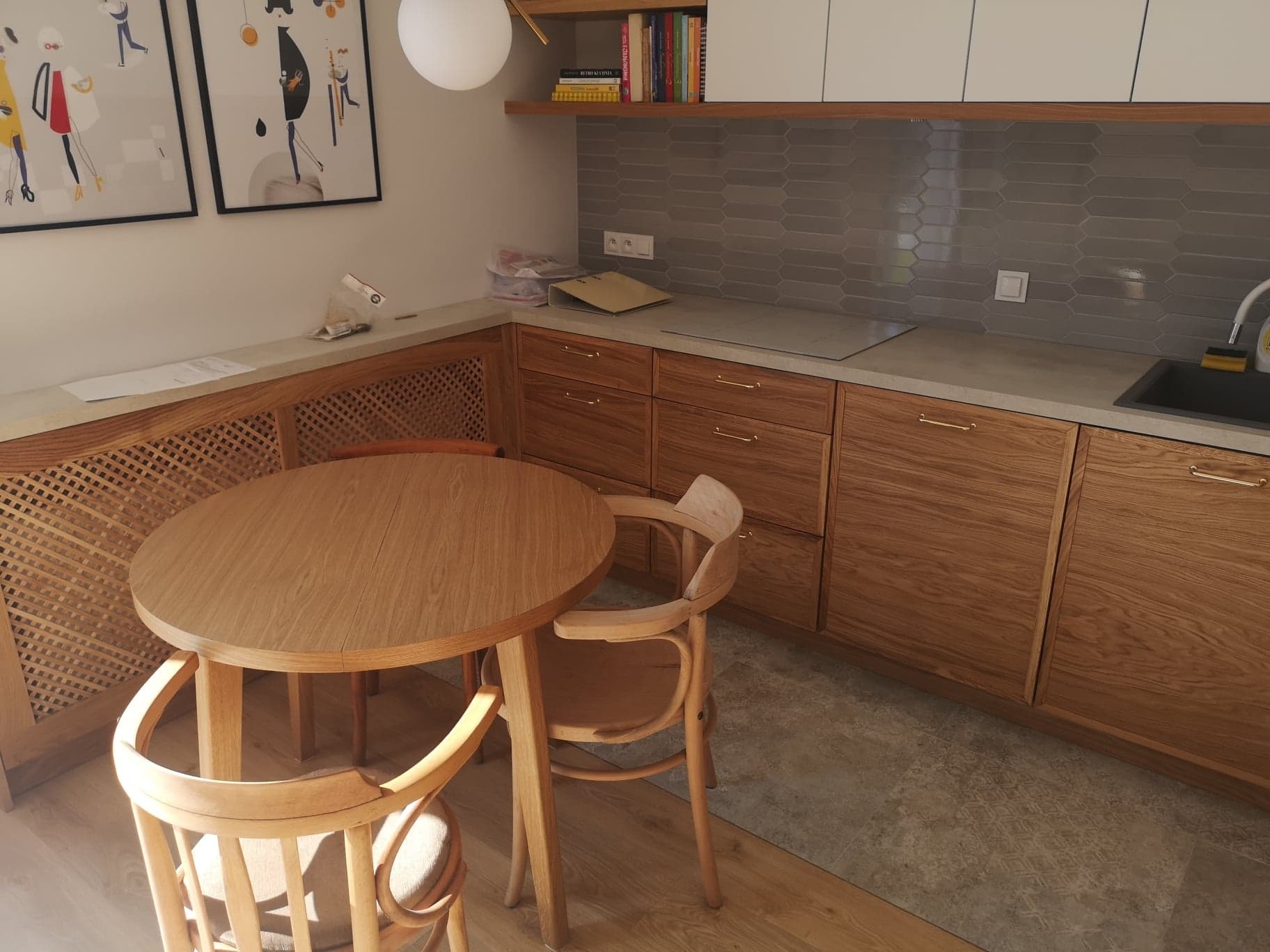 Drewniana zabudowa kuchni z okrągłym stołem i krzesłami w jasnym wnętrzu, szare płytki na ścianie między szafkami, obrazy na ścianie.