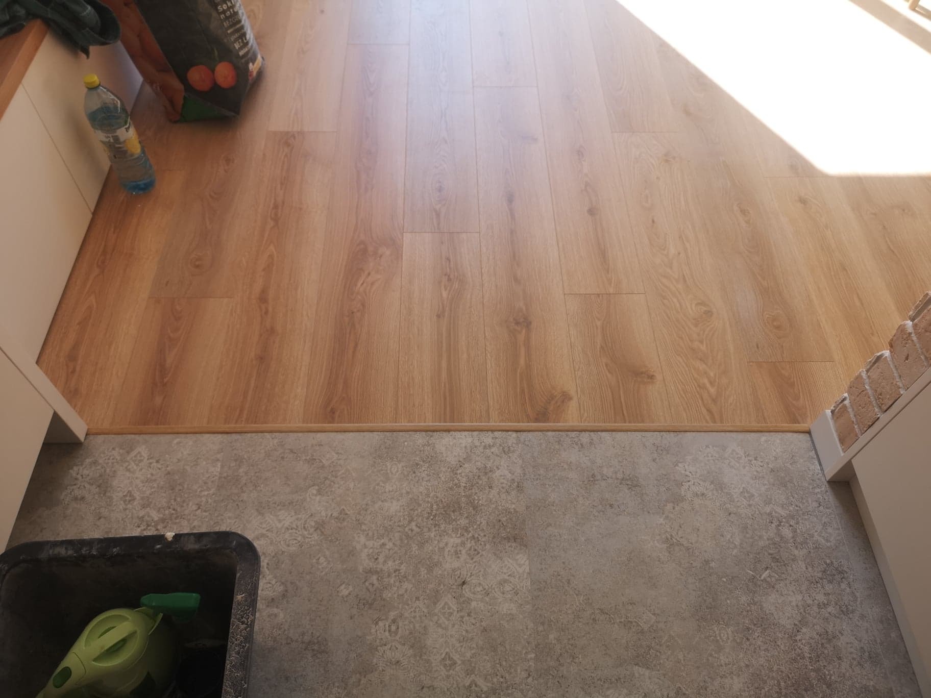 Ułożona podłoga z paneli drewnopodobnych przechodząca w płytki podłogowe z wzorem, widoczny fragment ściany z cegły i wiadro z akcesoriami do sprzątania.