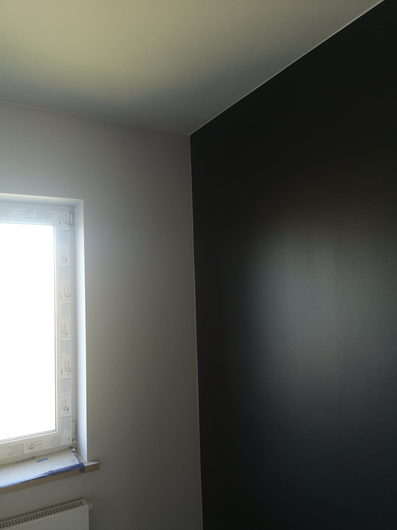 Pomalowane na czarno ściany w rogu pokoju, widoczne białe okno z plastikową ramą i zabezpieczonym parapetem oraz biały grzejnik pod nim.