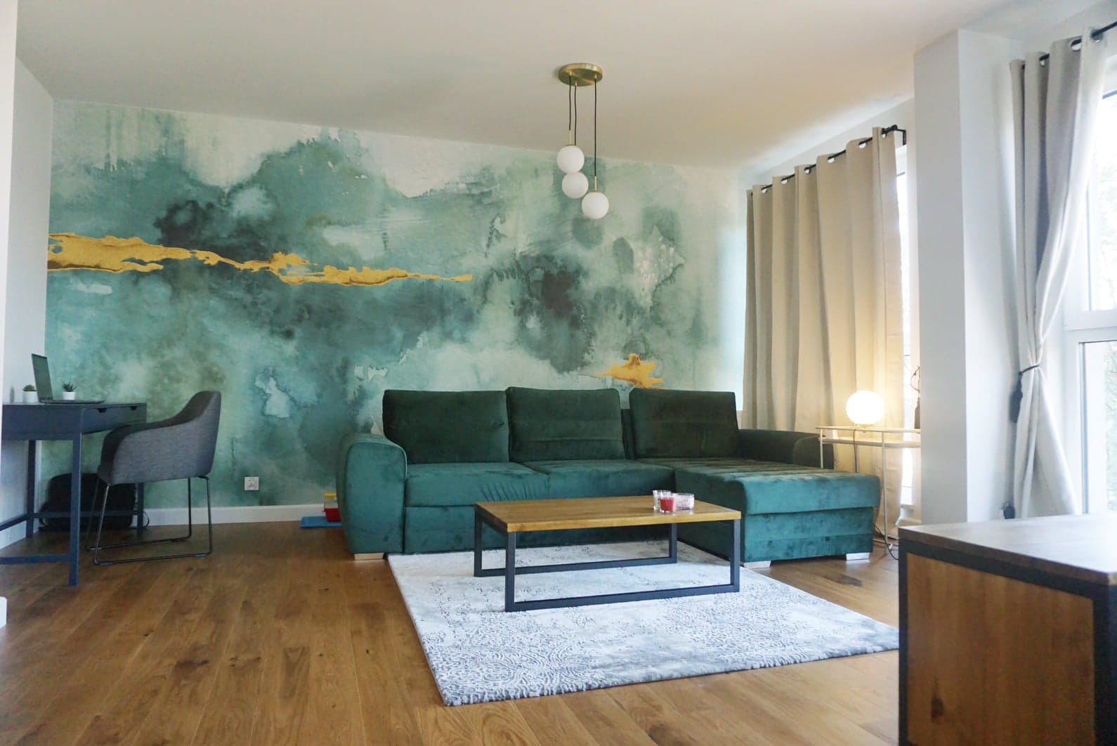 Nowoczesny salon z zieloną sofą narożną, drewnianym stolikiem kawowym, wzorzystą tapetą w odcieniach zieleni i złota oraz drewnianą podłogą