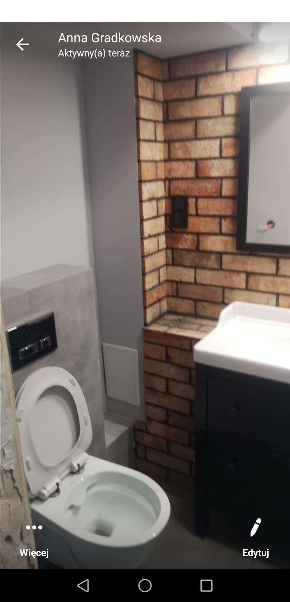 Ukończona łazienka z toaletą z otwartą klapą, umywalką na szafce w stylu retro i ścianą wykończoną cegłą.