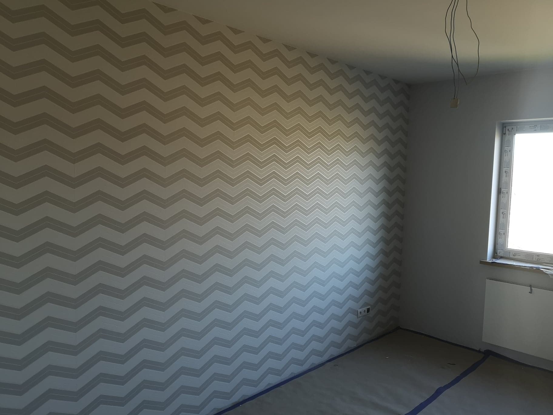 Pokój po tapetowaniu w zygzaki, widoczne wystające kable, okno i zabezpieczona podłoga.