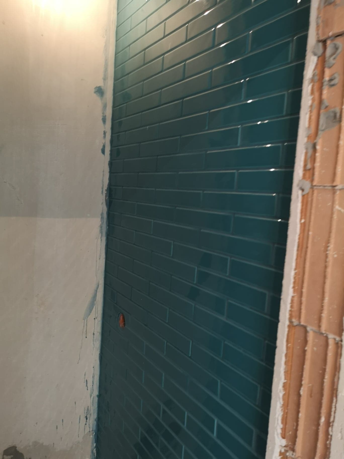 Ściana w trakcie układania płytek w odcieniach turkusu w układzie cegiełki, widoczny fragment surowej ściany i narożnik z cegieł.