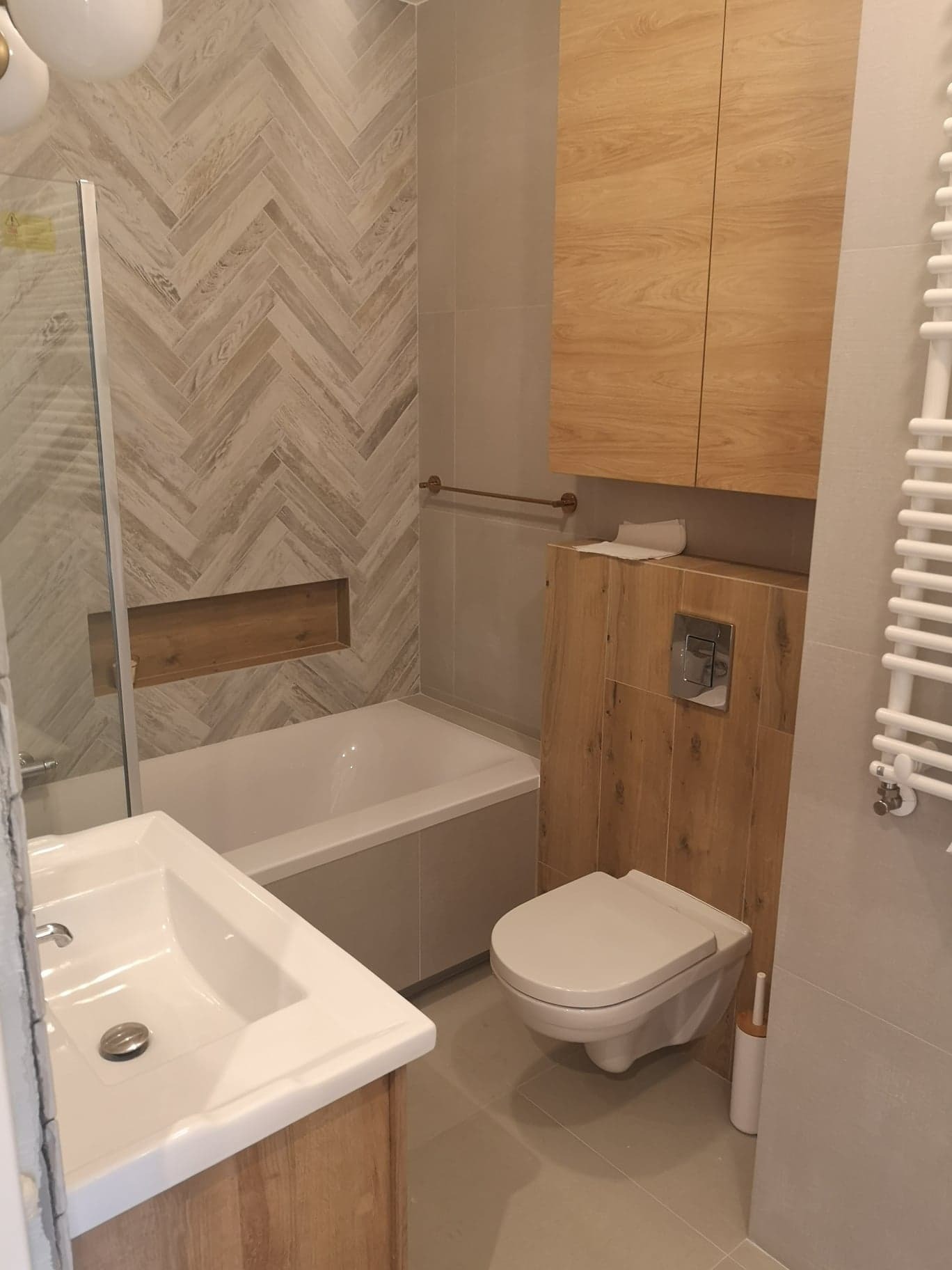 Nowoczesna łazienka z drewnopodobnymi płytkami ułożonymi w jodełkę na ścianie przy wannie, zabudowana szafka nad toaletą w tym samym stylu, biała umywalka z szafką, grzejnik drabinkowy...