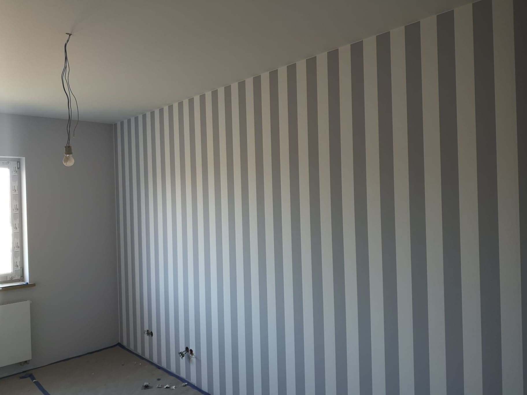 Ściana w pokoju po świeżym malowaniu w biało-szare pionowe pasy, widoczne niedokończone gniazdka elektryczne i wisząca żarówka bez klosza.