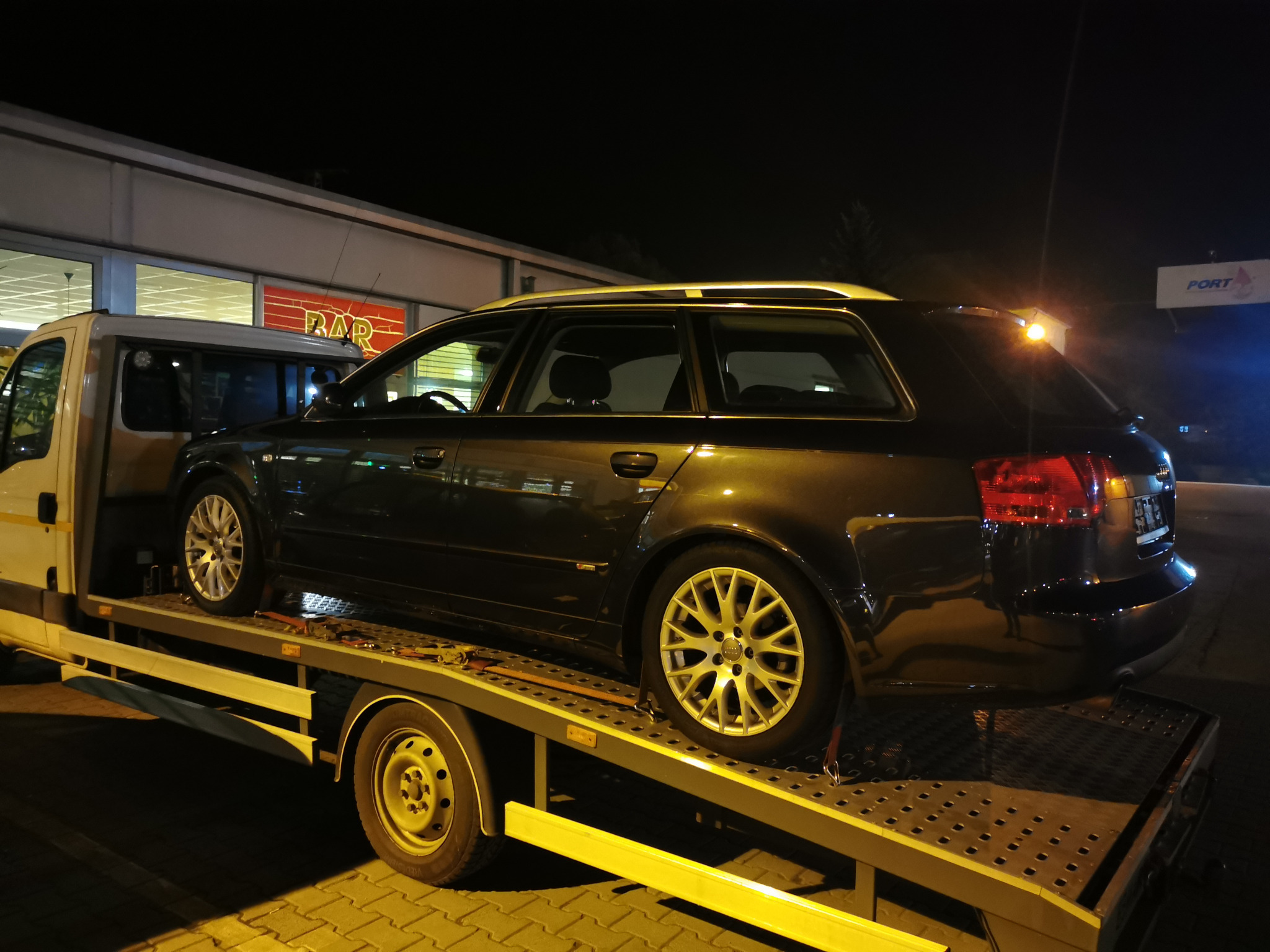 Czarny samochód kombi marki Audi zabezpieczony pasami transportowymi na platformie lawety w nocy, widoczne światła budynku w tle.