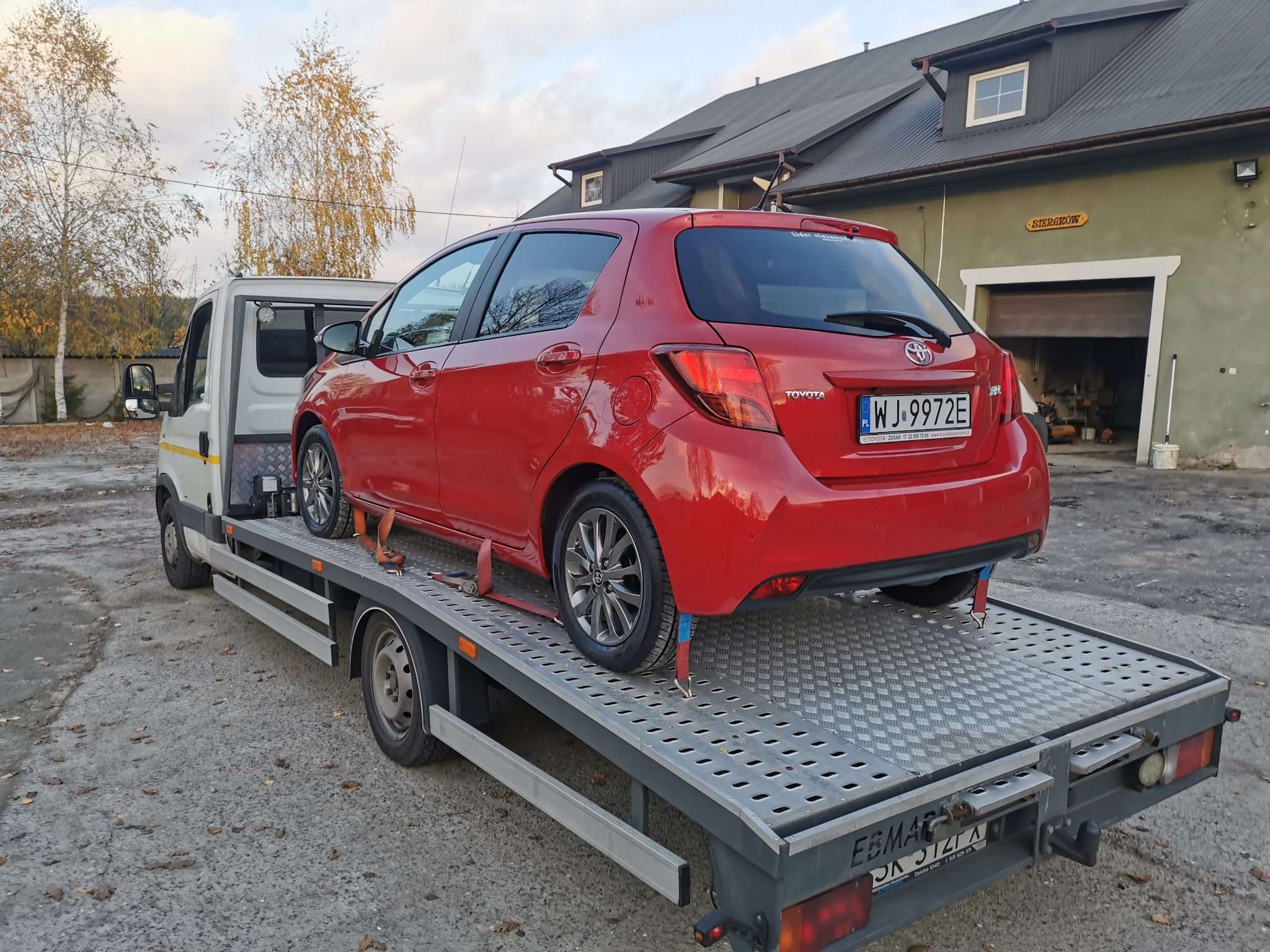 Czerwona Toyota Yaris zabezpieczona pasami na platformie lawety z metalowym podłożem w trakcie transportu, widoczny numer rejestracyjny WJ 9972E i budynek w tle.