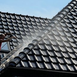 Roofers Folding Group - Pracownik w czerwonej koszulce czyści czarny dach z dachówki ceramicznej za pomocą myjki ciśnieniowej, stojąc na aluminiowej drabinie opierającej się o okno dachowe.
