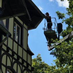 Roofers Folding Group - Pracownik na podnośniku koszowym, w czapce i roboczych ubraniach, dokonuje inspekcji lub drobnych napraw przy okapie dachu starego budynku z widoczną konstrukcją szachulcową i metalowymi rynnami...